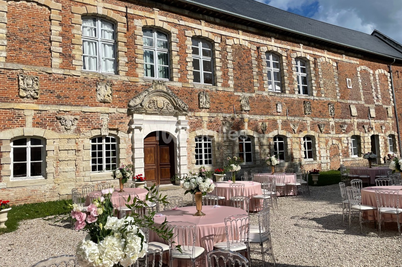 Cour d'un bâtiment en briques et pierres, aménagée avec des tables décorées de nappes roses et de bouquets de fleurs.