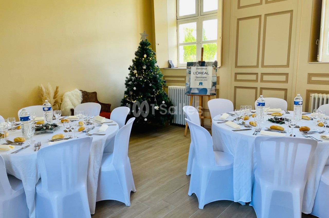 Salle décorée pour un repas, avec tables rondes dressées, nappes blanches et un sapin de Noël illuminé.