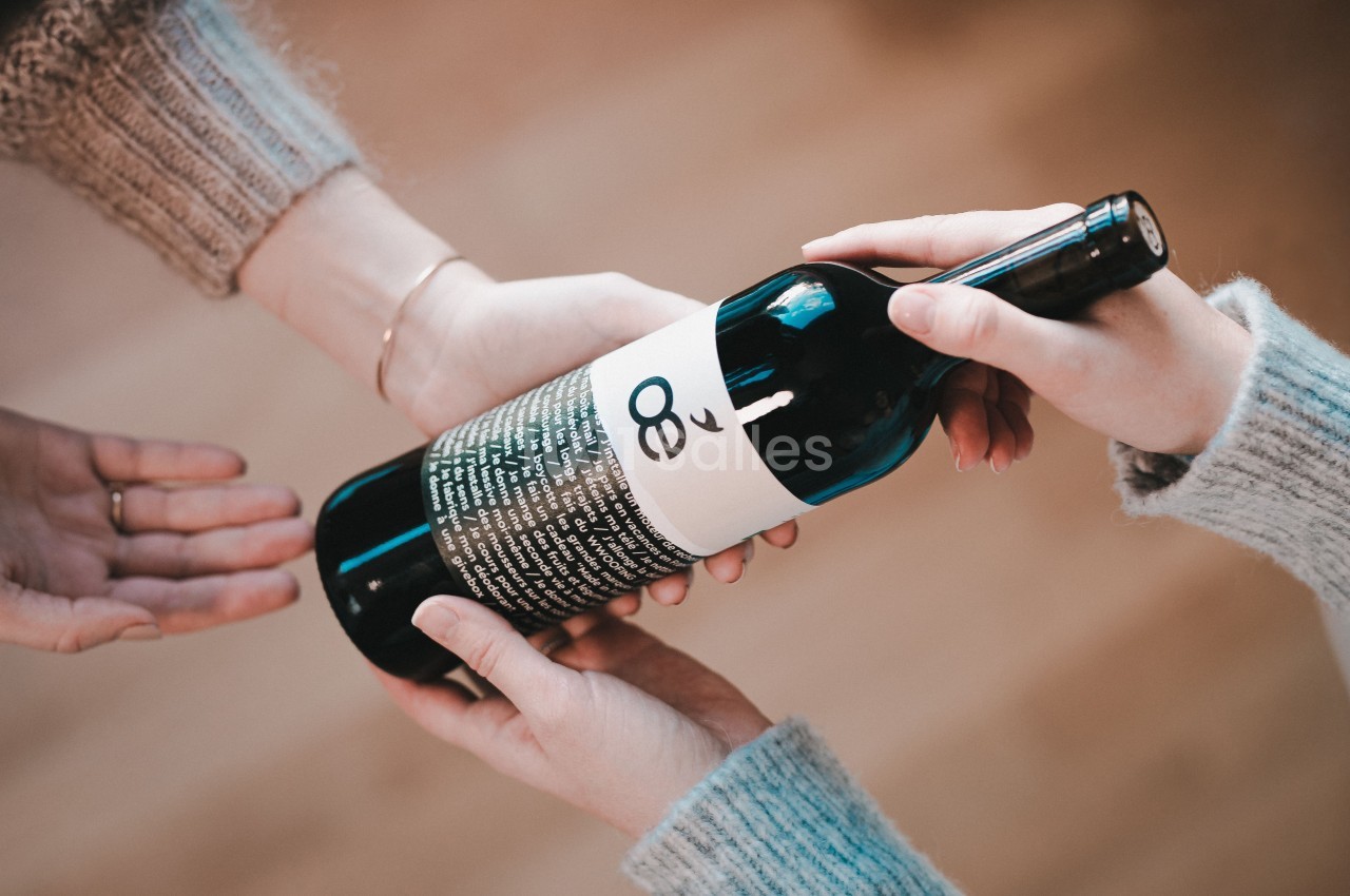 Deux mains tendent une bouteille de vin à une troisième main dans un environnement intérieur lumineux.