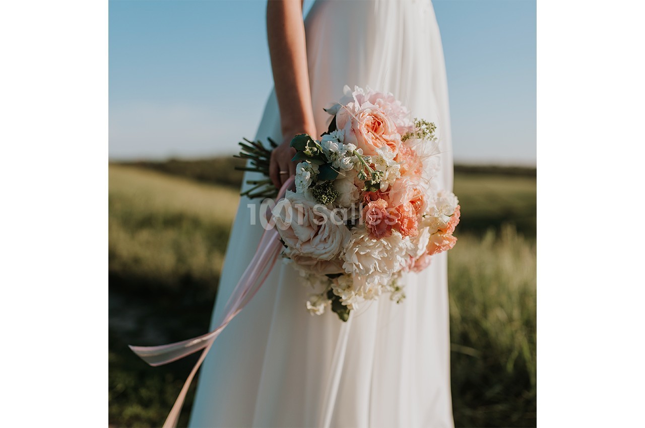 Une mariée en robe blanche tient un bouquet de fleurs pastel dans un champ sous un ciel dégagé.