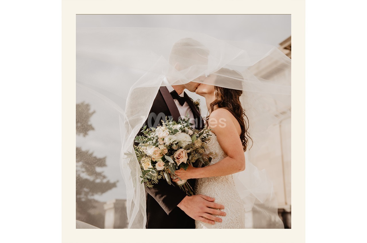 Un couple en tenue de mariage s'embrasse sous un voile, tenant un bouquet de fleurs.