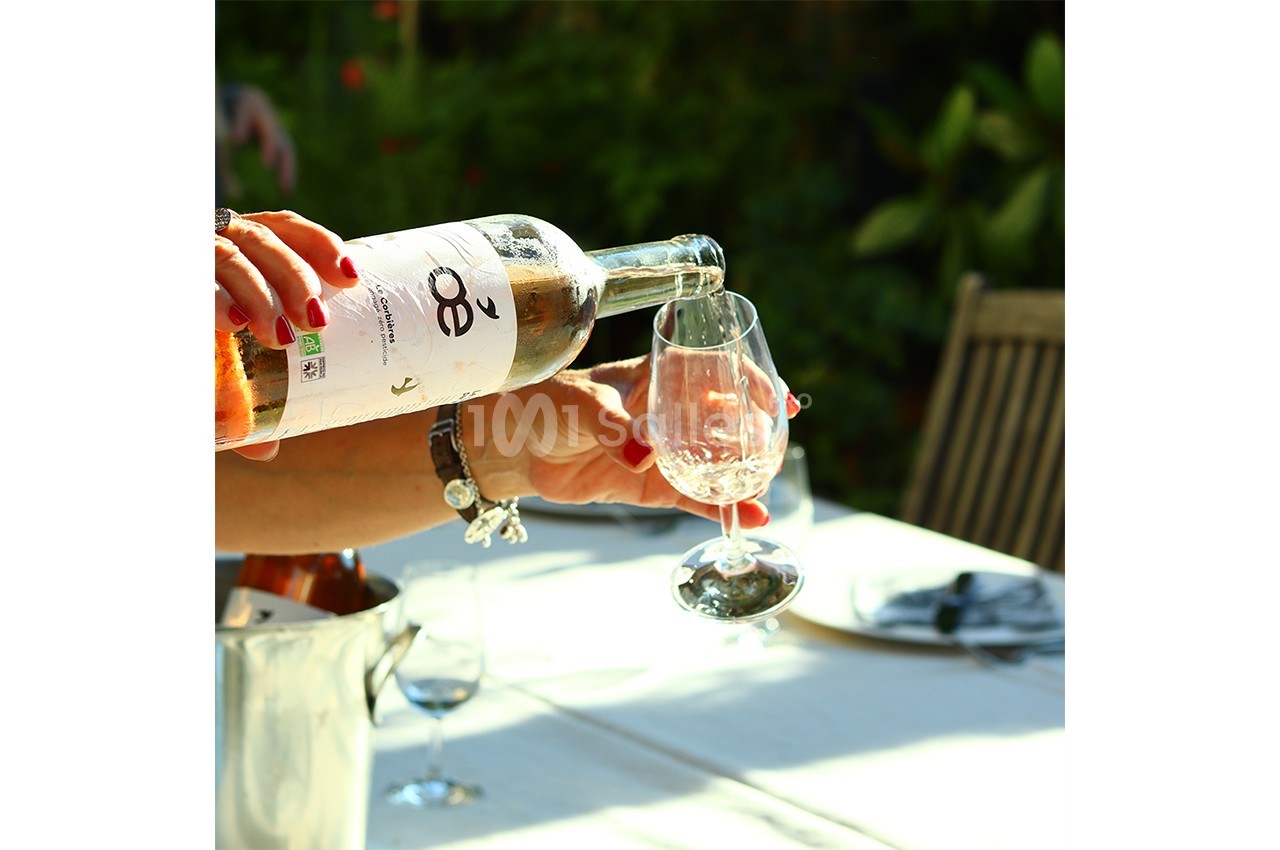Une personne verse du vin rosé dans un verre sur une table dressée en extérieur, entourée de verdure.