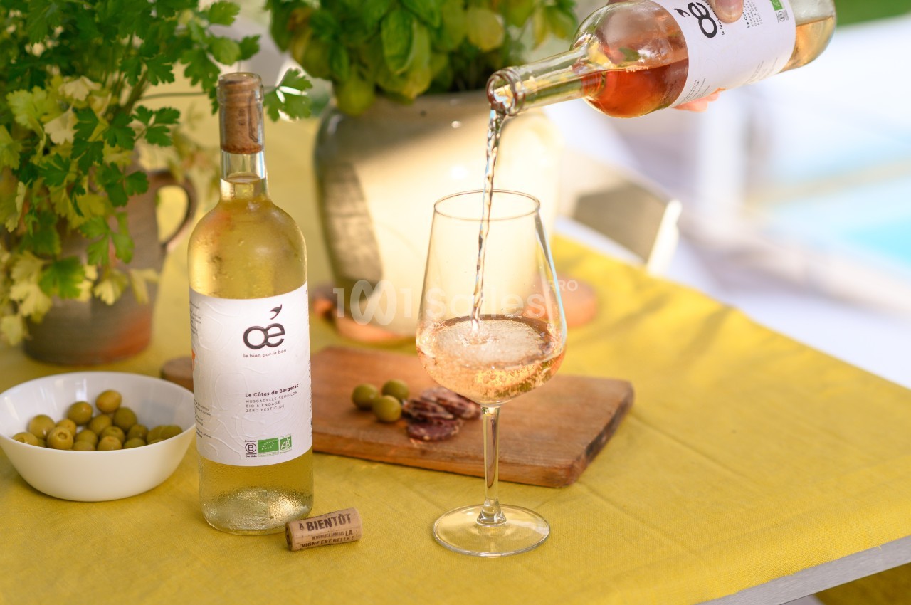 Une bouteille de vin blanc bio, un verre en cours de remplissage, des olives et des herbes sur une table jaune.