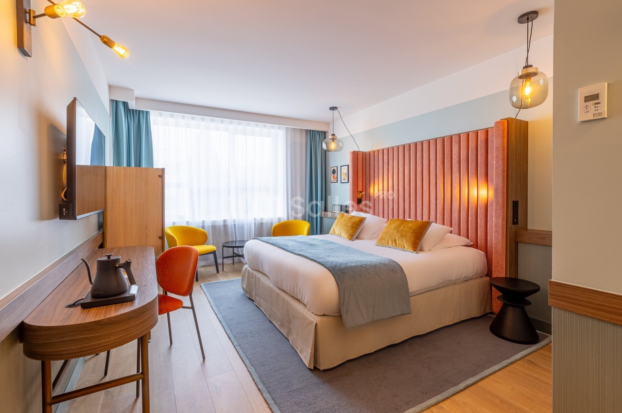 Chambre d'hôtel lumineuse avec un lit double, un bureau, des fauteuils colorés et une grande fenêtre avec rideaux.