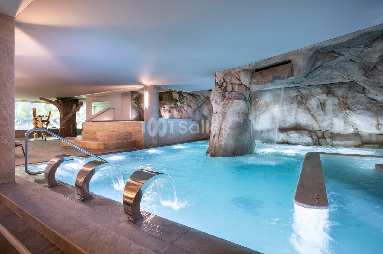 Piscine intérieure avec jets d'eau, cascade artificielle et décor rocheux dans un espace lumineux.