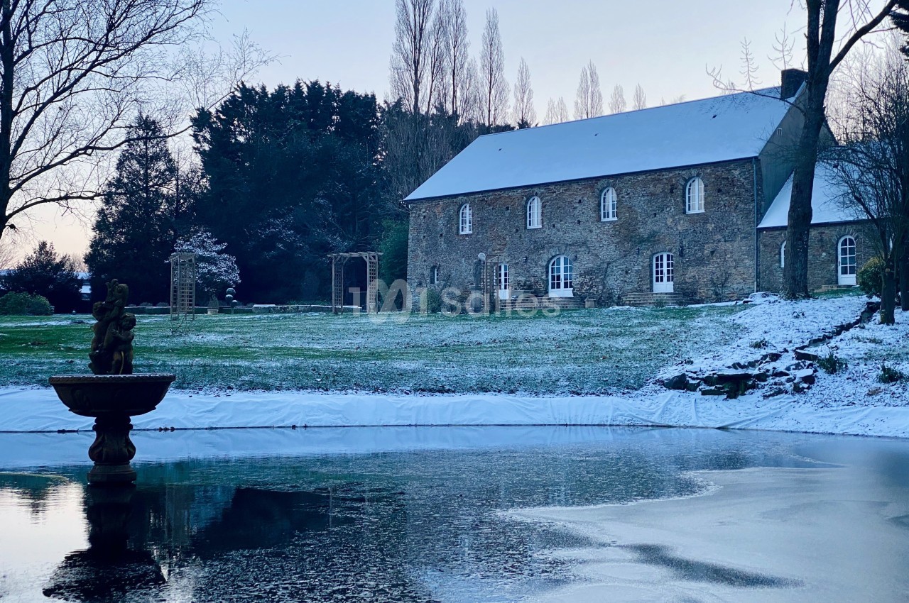Manoir en pierre entouré d'un jardin enneigé avec un bassin gelé et une fontaine au premier plan.