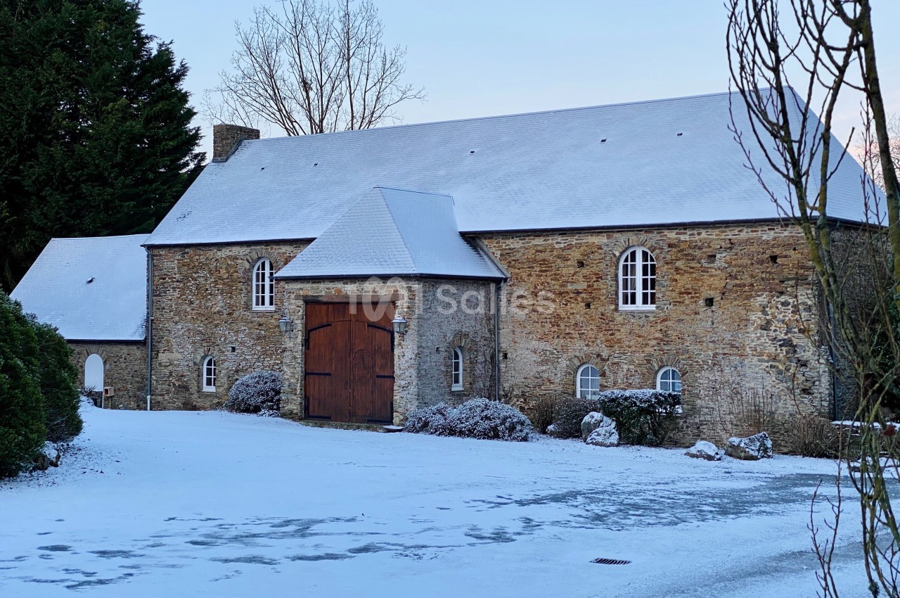 Bâtisse en pierre avec toit enneigé, grande porte en bois, entourée d'arbres et d'un sol recouvert de neige.