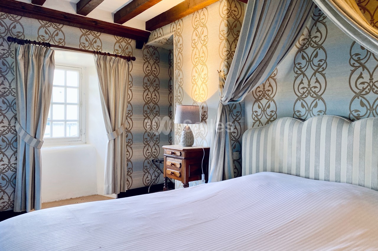 Chambre lumineuse avec lit à baldaquin, murs ornés de motifs, poutres apparentes et fenêtre encadrée de rideaux.