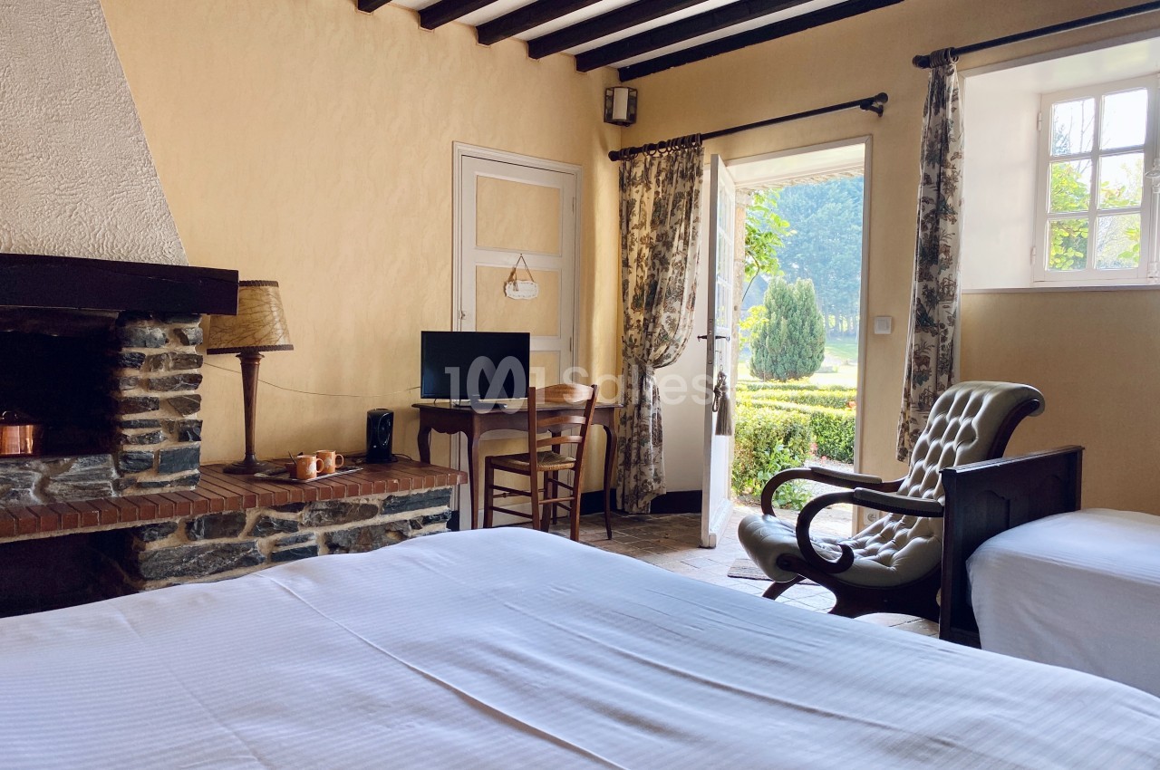 Chambre lumineuse avec lit, cheminée en pierre, bureau, fauteuil et vue sur un jardin à travers une porte ouverte.