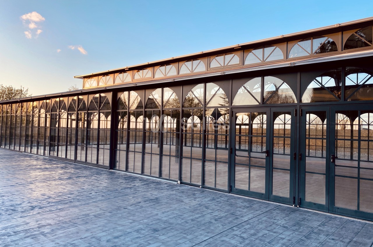 Grande serre en verre avec structure métallique et portes vitrées, reflétant le ciel au coucher du soleil.
