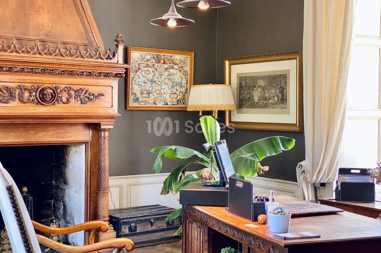 Bureau élégant avec cheminée en bois sculpté, plantes, cartes encadrées et lumière naturelle entrant par une fenêtre.