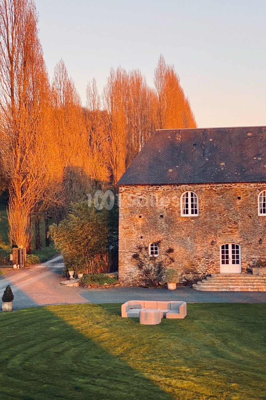 Maison en pierre éclairée par la lumière du coucher de soleil, entourée d'arbres et d'une pelouse bien entretenue.