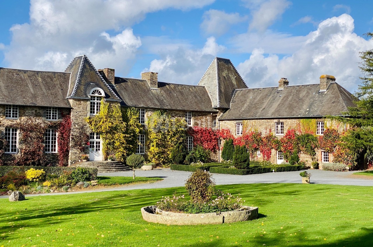 Manoir en pierre entouré de vignes rouges et jaunes, avec pelouse et parterre fleuri au premier plan.
