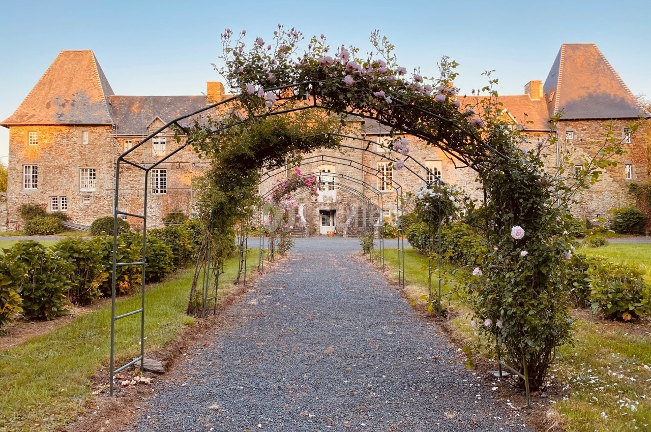 Allée bordée d'arceaux fleuris menant à un bâtiment en pierre sous un ciel dégagé.