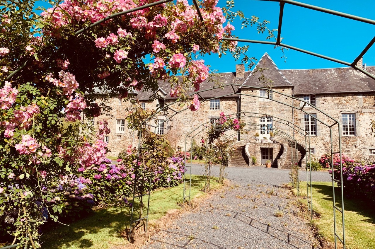 Allée fleurie avec des arches métalliques menant à un bâtiment en pierre sous un ciel bleu.