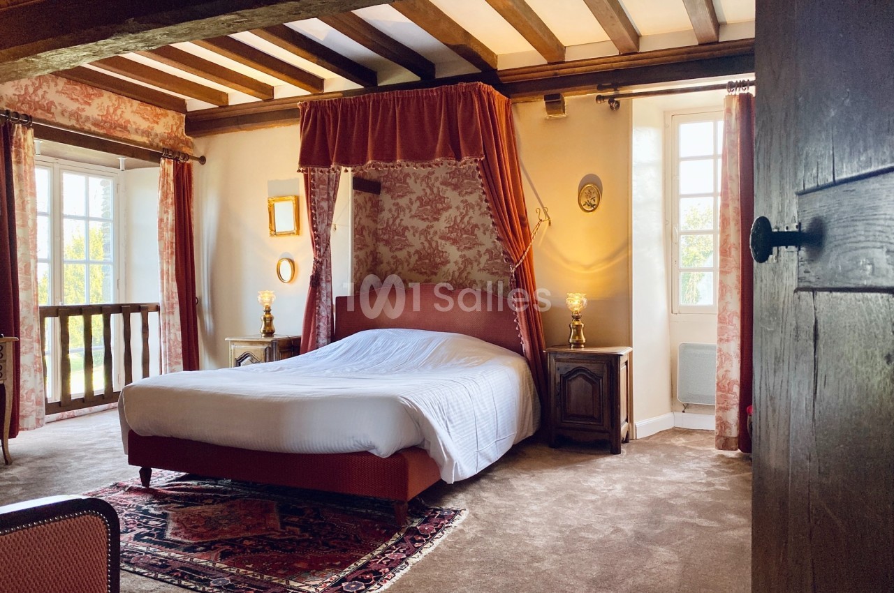 Chambre lumineuse avec lit à baldaquin, tapis rouge, poutres apparentes et fenêtres donnant sur un extérieur verdoyant.