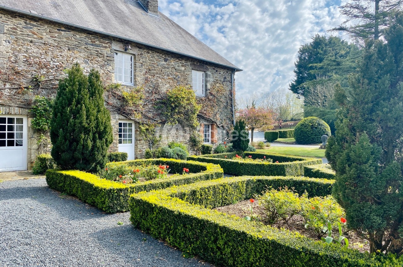 Maison en pierre avec jardin structuré de haies taillées, parsemé de fleurs, sous un ciel partiellement nuageux.