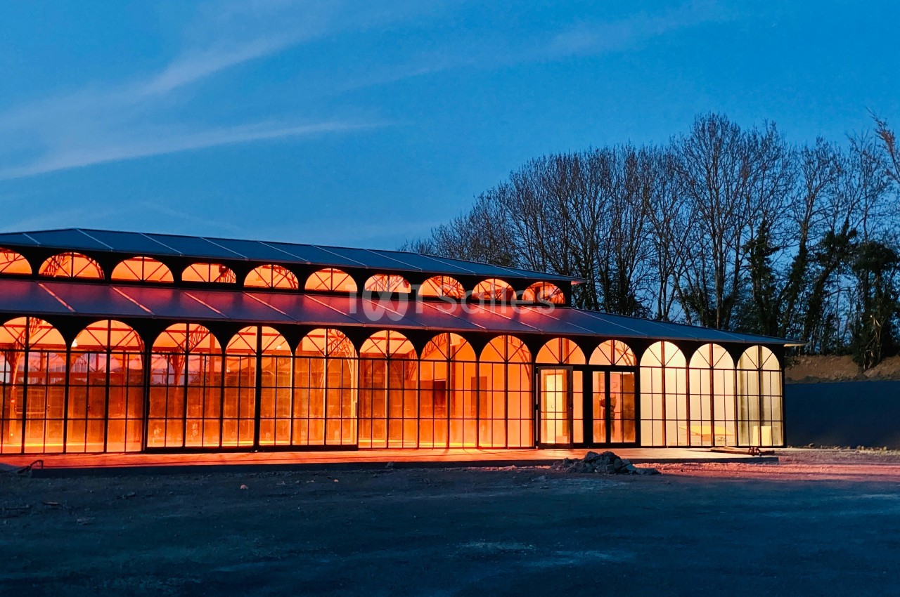 Bâtiment vitré illuminé en orange au crépuscule, entouré d'arbres et d'un sol dégagé.