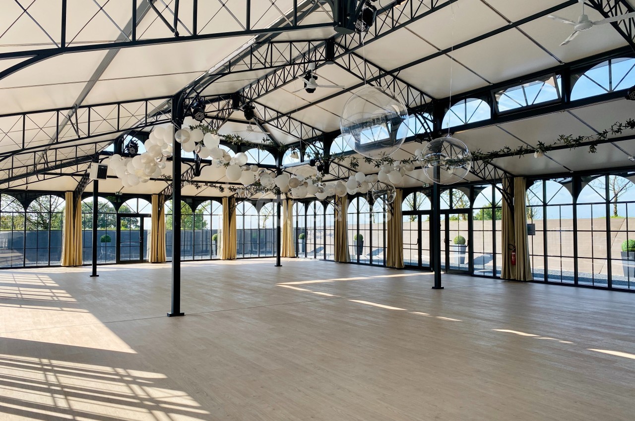 Salle spacieuse avec toit en verrière, structures métalliques noires et décorations suspendues, baignée de lumière naturelle.