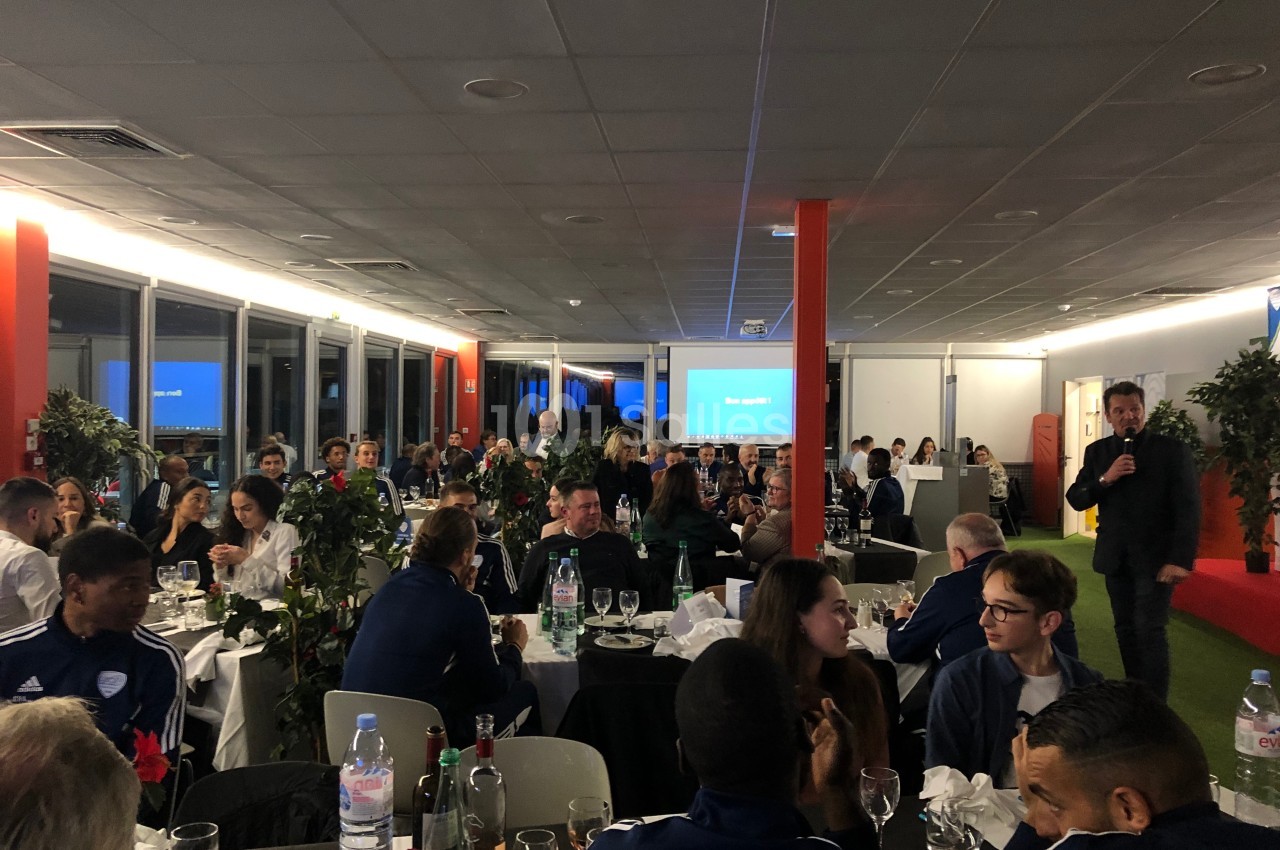 Salle remplie de personnes assises à des tables, lors d'un événement avec un intervenant debout et un écran en arrière-plan.