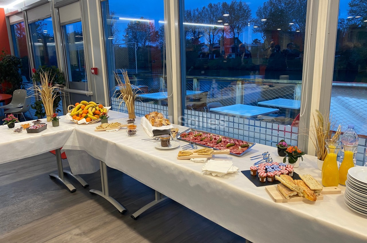 Buffet dressé avec fruits, viennoiseries, jus et mets variés, dans une salle lumineuse avec vue sur l'extérieur.