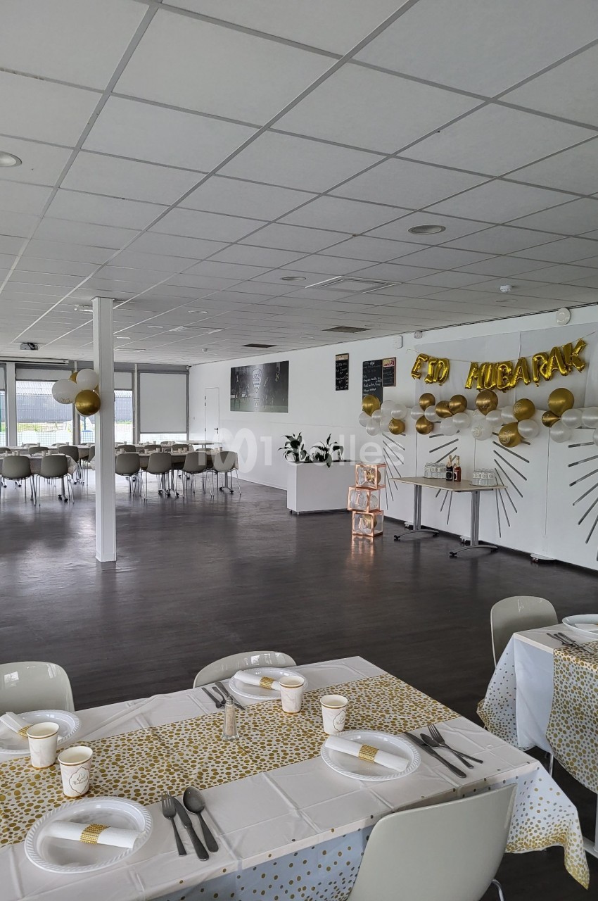Salle décorée pour une fête avec des tables dressées, des ballons dorés et une bannière ’Eid Mubarak’.