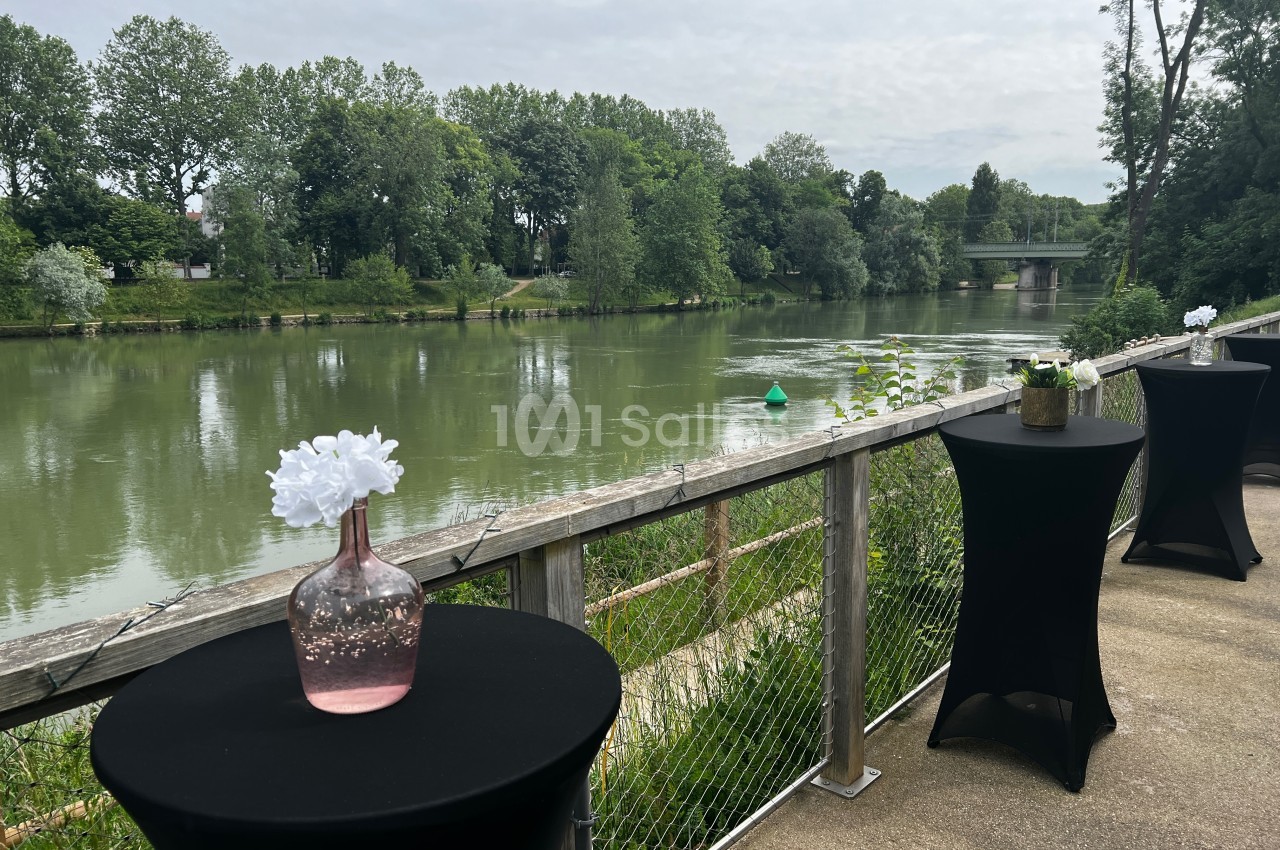 Tables hautes avec nappes noires et vases fleuris sur une terrasse surplombant une rivière bordée d'arbres.
