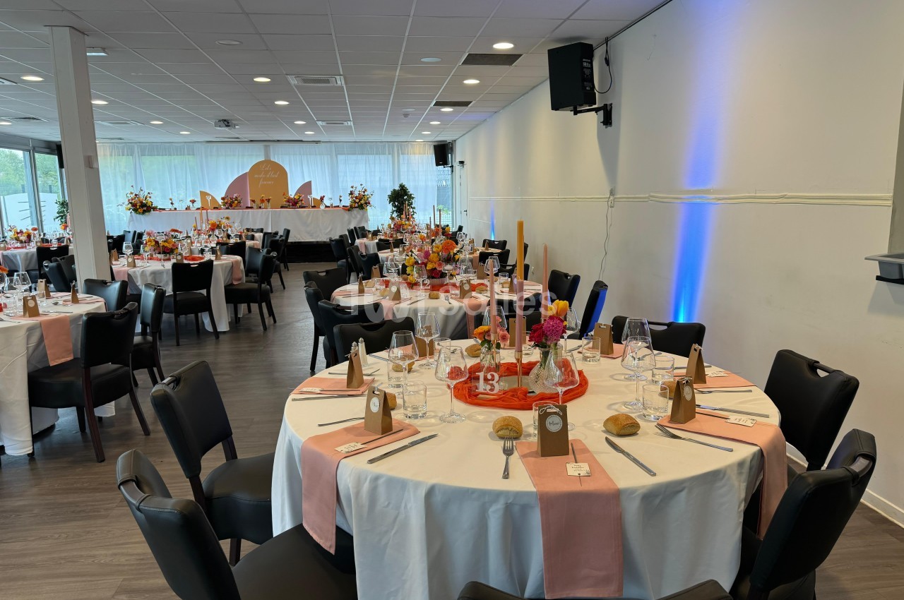 Salle de réception décorée avec des tables rondes dressées, nappes blanches, chemins de table roses et arrangements floraux.