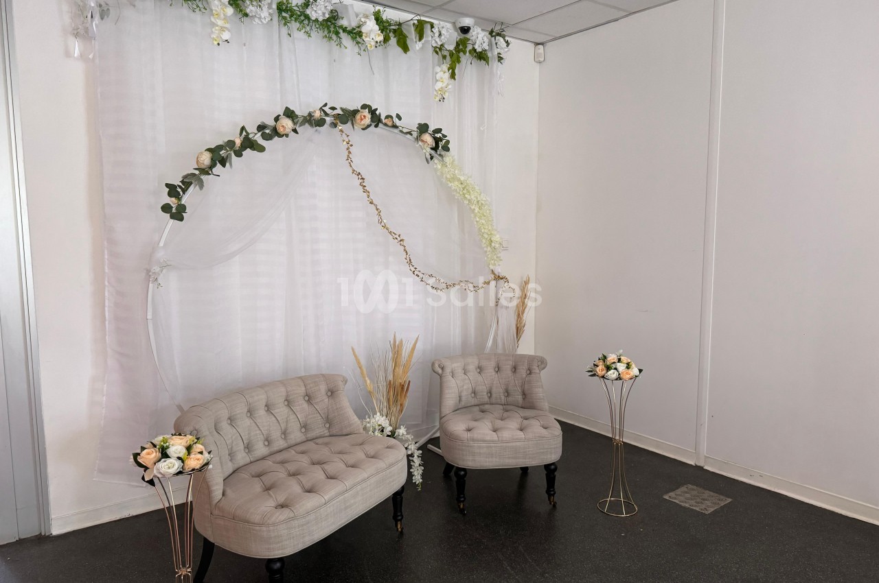 Deux fauteuils beiges devant un rideau blanc décoré de fleurs et de feuillages, avec des bouquets sur des supports dorés.
