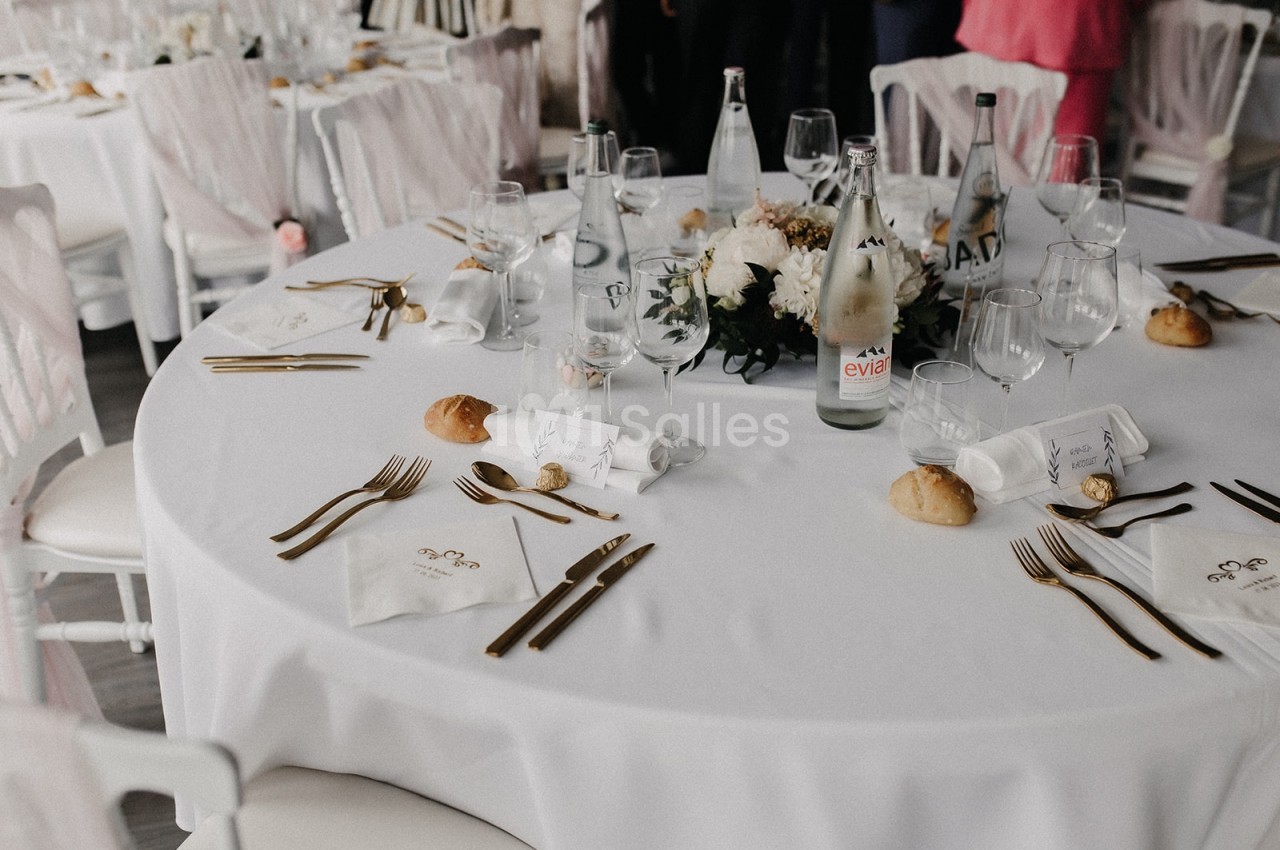 Table ronde dressée pour un repas, avec nappes blanches, couverts dorés, verres, fleurs et petits pains.