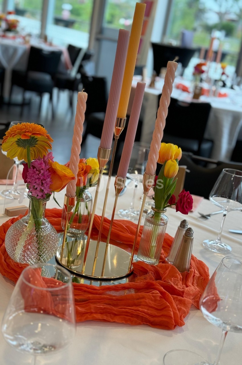Centre de table avec bougies torsadées, fleurs colorées et nappe orange, entouré de verres et couverts.