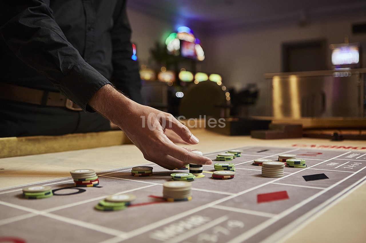 Un croupier place des jetons sur une table de roulette dans un casino, avec des machines à sous en arrière-plan.