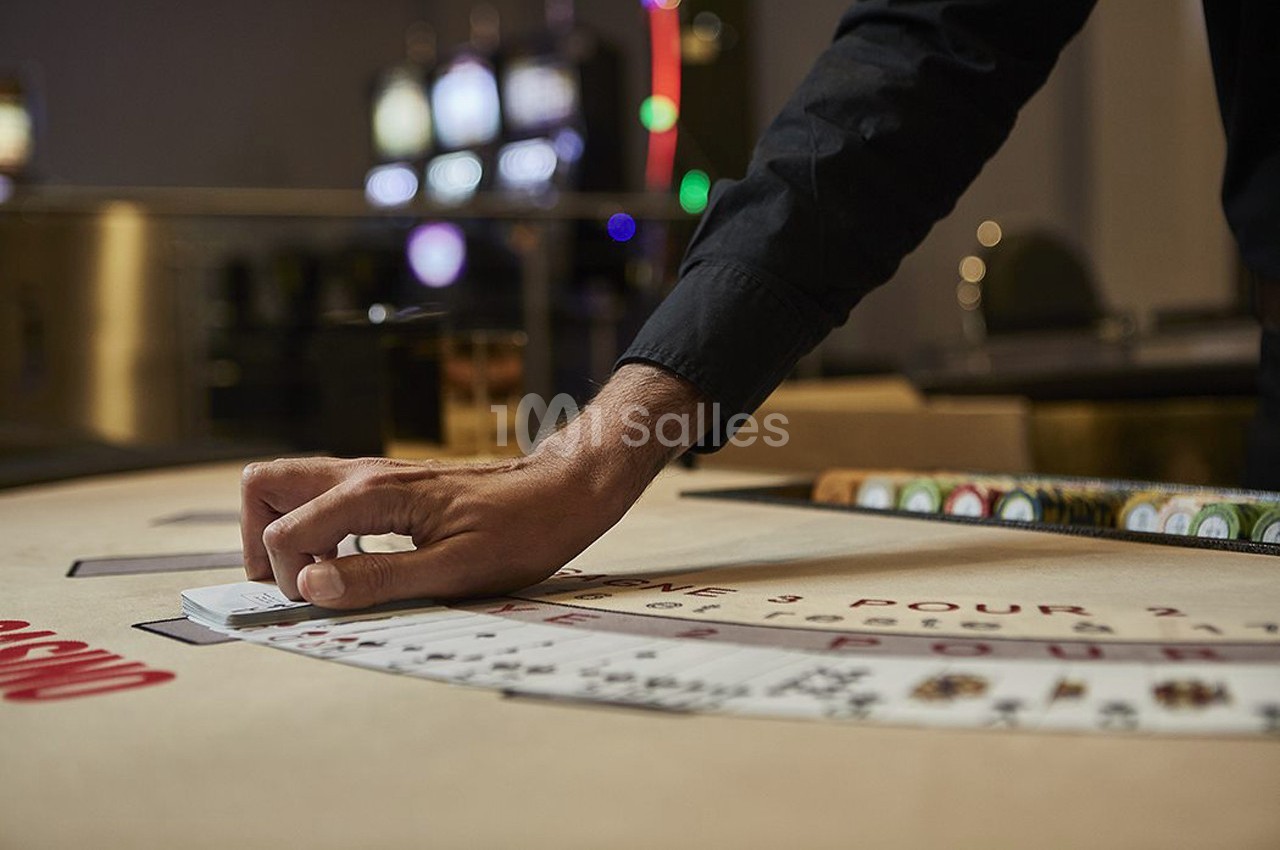 Main distribuant des cartes sur une table de jeu de casino avec des machines à sous en arrière-plan flou.