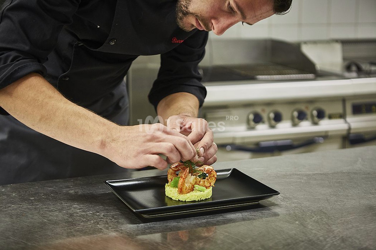 Un chef dispose soigneusement des crevettes sur un plat noir dans une cuisine professionnelle.
