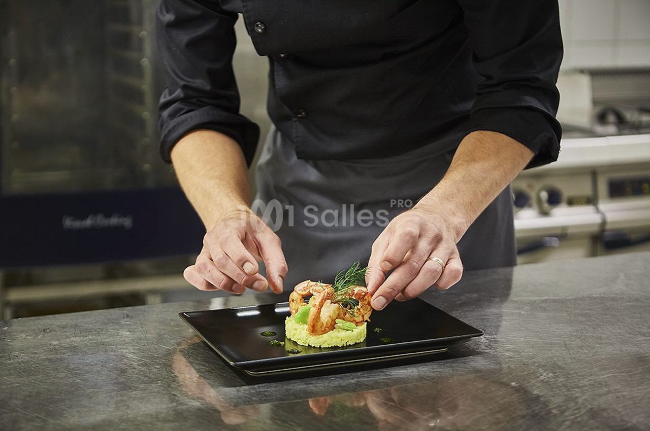 Un chef dispose délicatement des crevettes sur un plat noir dans une cuisine professionnelle.