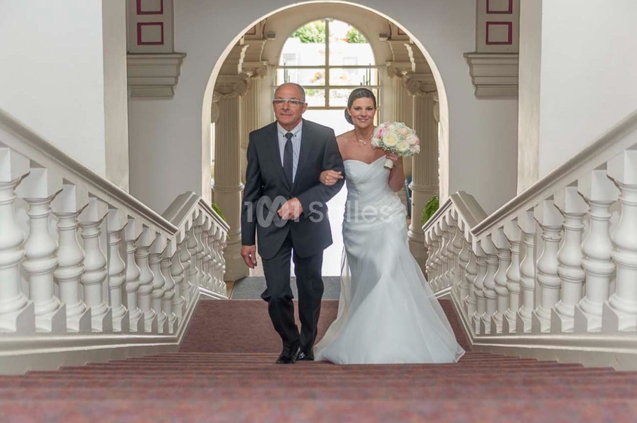 Un homme en costume accompagne une mariée en robe blanche montant un escalier dans un bâtiment lumineux.