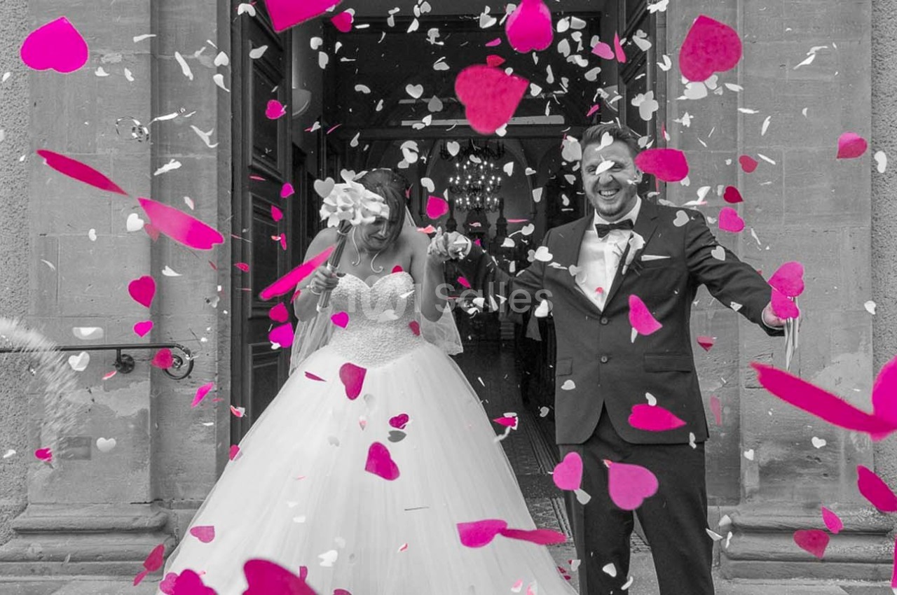 Un couple souriant en tenue de mariage sort d'un bâtiment sous une pluie de confettis roses et blancs.