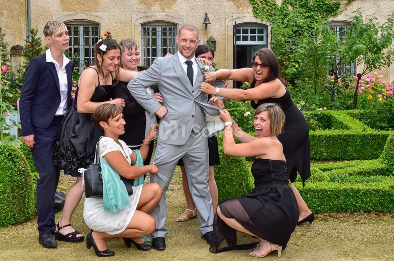 Un homme en costume gris entouré de femmes en tenue de soirée dans un jardin devant un bâtiment en pierre.