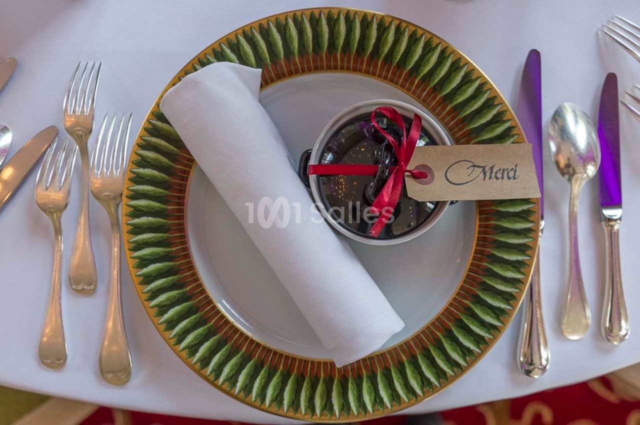 Assiette décorative avec serviette blanche roulée et petit pot cadeau avec étiquette ’Merci’, entourée de couverts.