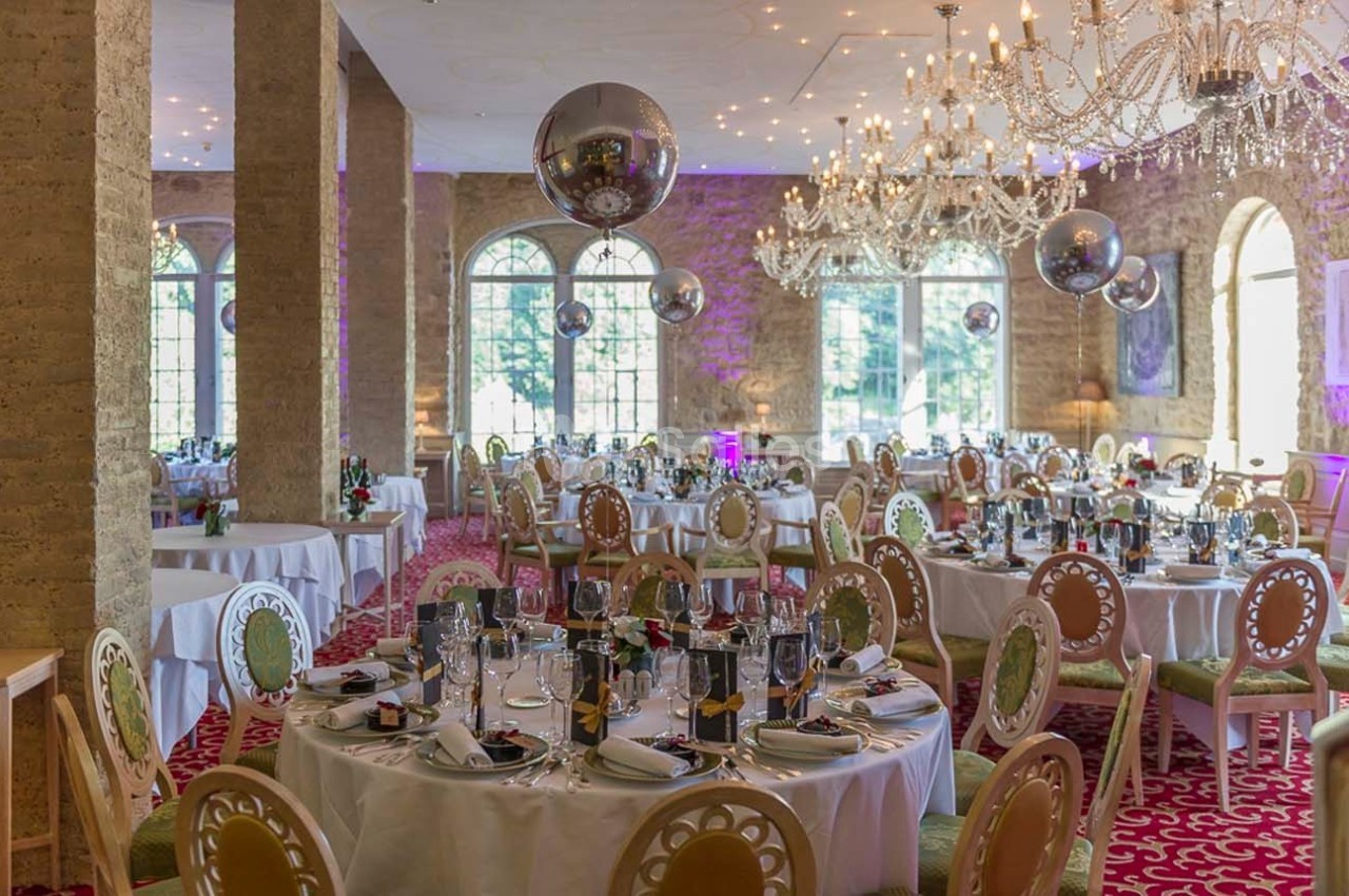 Salle de réception élégante avec tables rondes dressées, lustres en cristal et grandes baies vitrées.