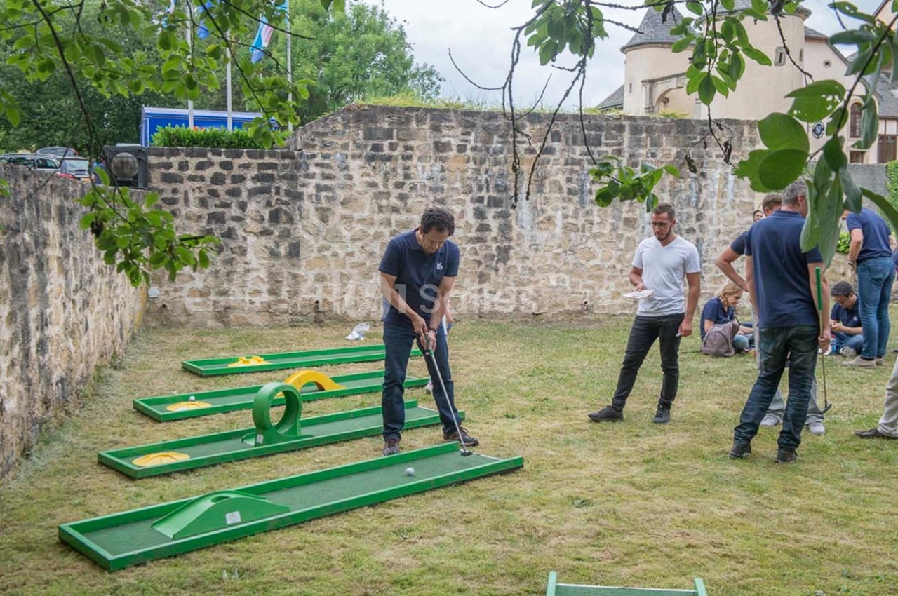 Un homme joue au mini-golf sur une pelouse avec des obstacles, entouré de plusieurs personnes.