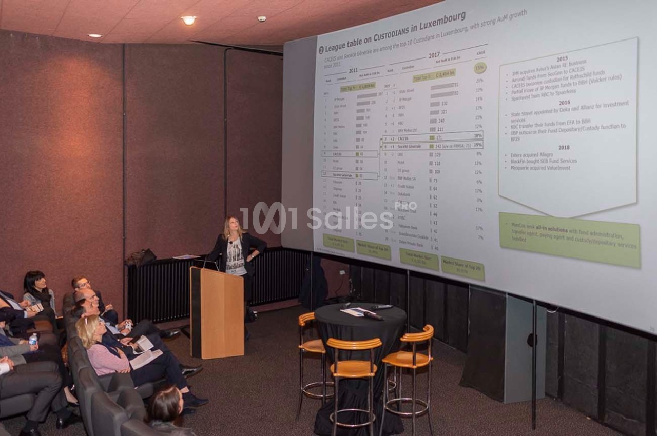 Une conférencière présente un tableau de données projeté sur un écran à un public assis dans une salle sombre.