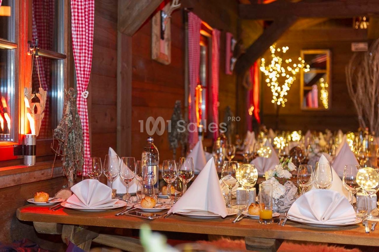 Table dressée dans un chalet en bois, décorée avec des nappes blanches, verres, bougies et lumières chaleureuses.
