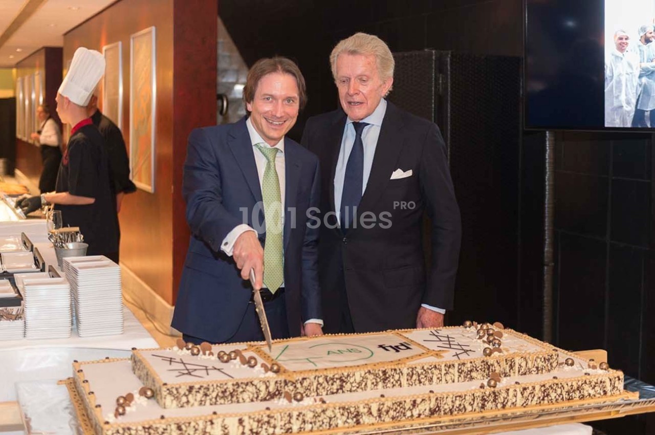 Deux hommes en costume coupent un grand gâteau décoré, dans un cadre formel avec un chef en arrière-plan.