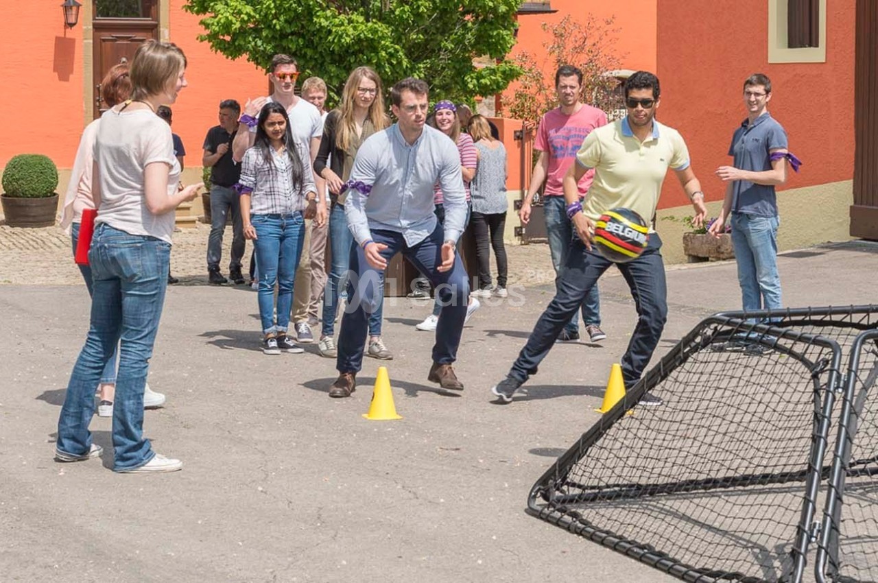 Des personnes participent à un jeu en plein air impliquant des cônes et un filet, dans une cour ensoleillée.