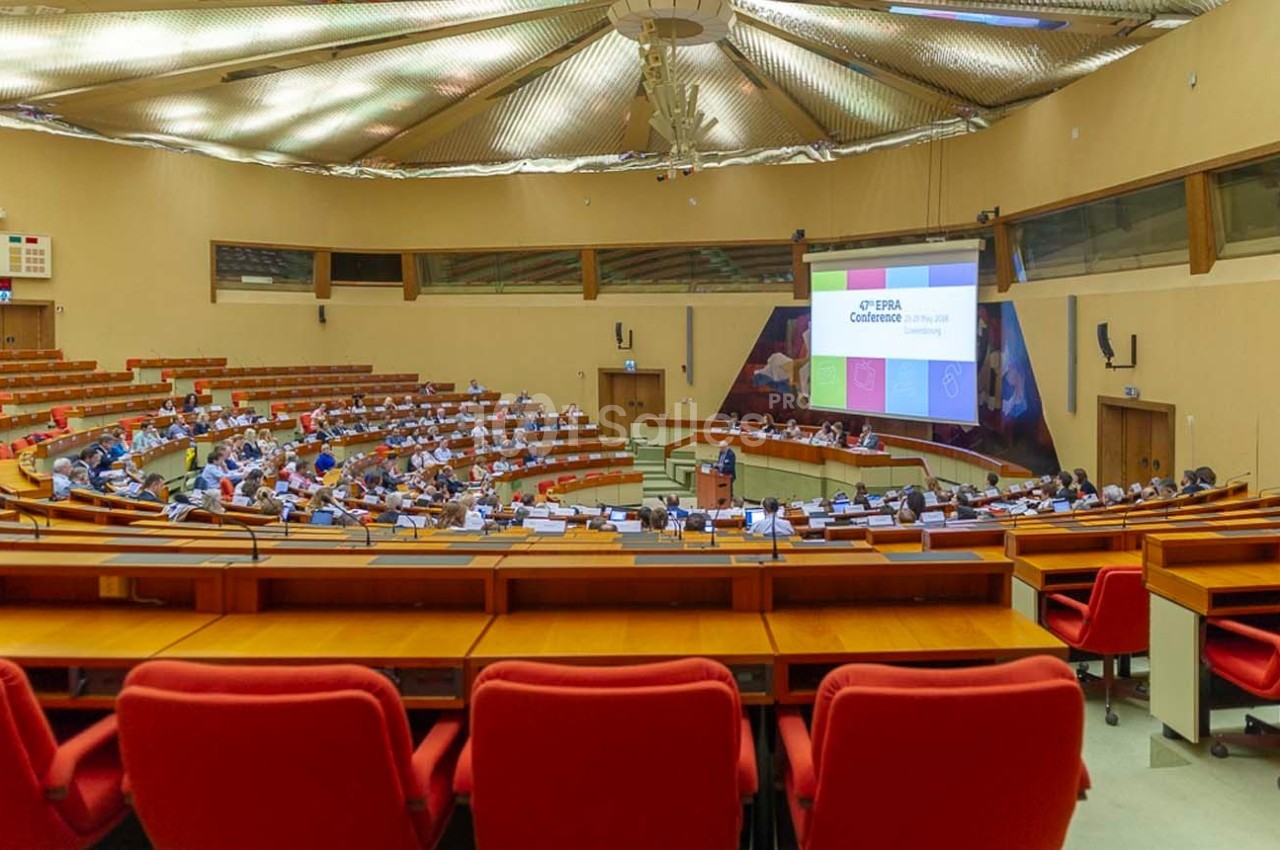 Salle de conférence avec des participants assis, un orateur debout et une présentation projetée à l'écran.
