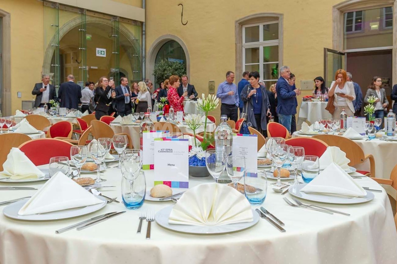 Salle de réception avec tables dressées, nappes blanches, verres et menus, invités debout en arrière-plan.