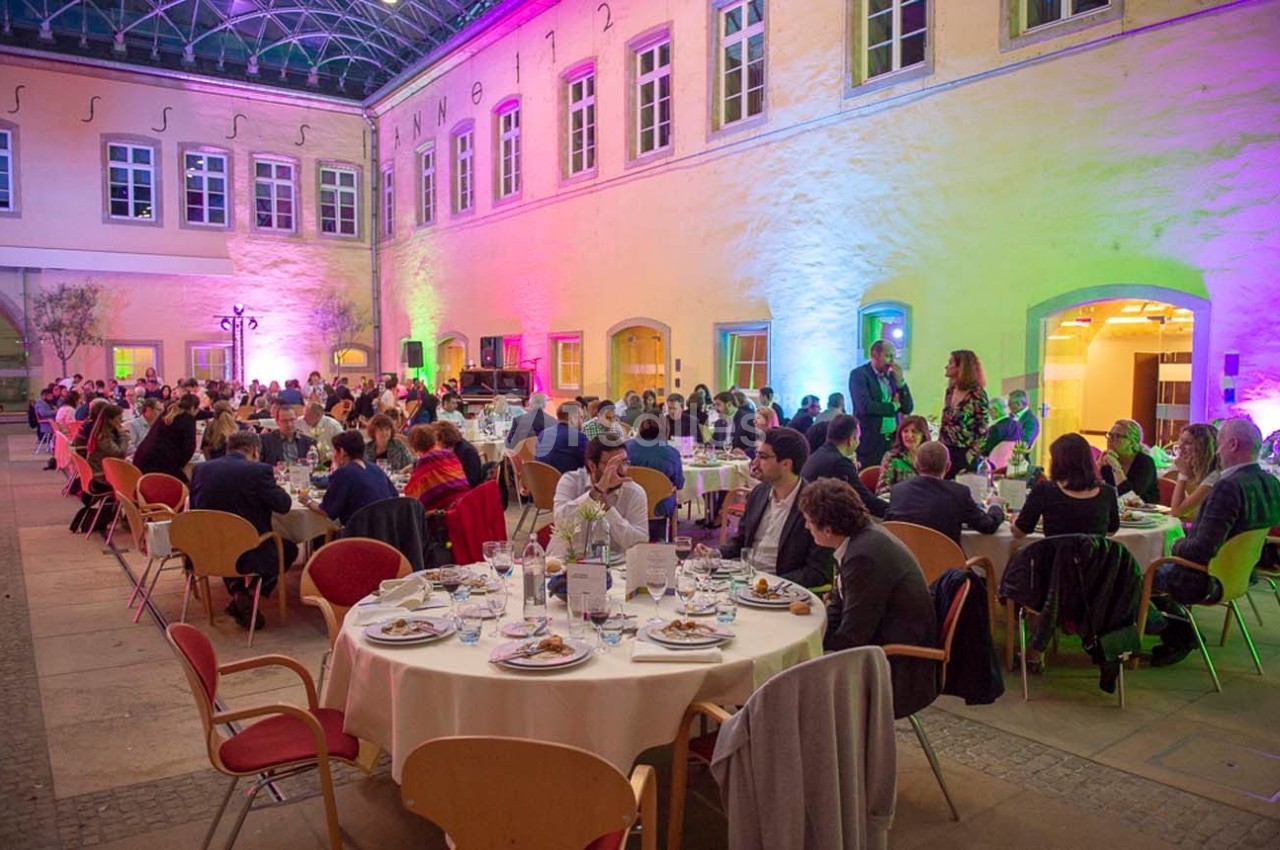 Des convives assis à des tables rondes dans une cour intérieure éclairée par des lumières colorées.