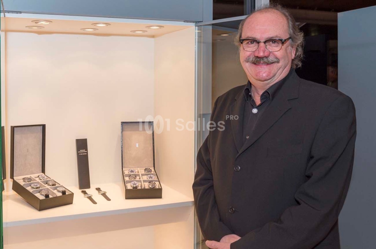 Un homme en costume se tient à côté d'une vitrine éclairée contenant des montres et leurs boîtes.