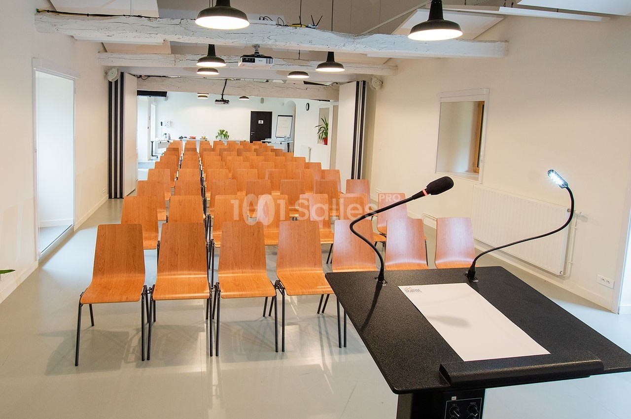Salle de conférence lumineuse avec rangées de chaises en bois et un pupitre équipé de micros au premier plan.
